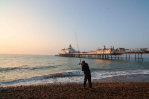 Early morning fisherman casts a line into the surf beside the onamental Victorian fun pier at Eastbourne, East Sussex at sunrise. Designed by naval architect Eugenius Birch, the famous seaside fun pier opened 13 June 1870